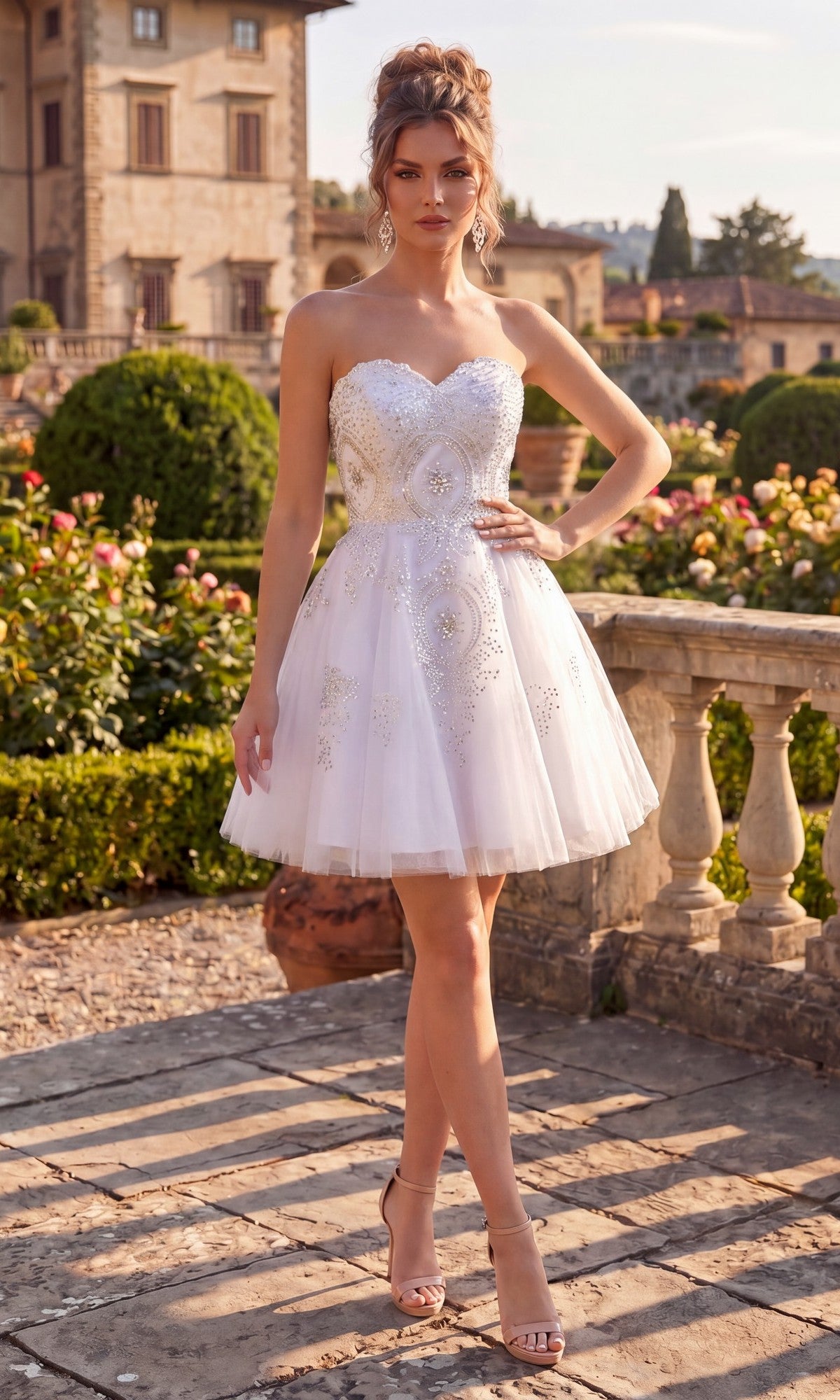 A woman wearing the Chicas C3350 Short Homecoming Dress in white, featuring a strapless fit-and-flare silhouette with beautiful embellishments, stands outdoors on a stone terrace with a lush garden and large house in the background. The elegant dress highlights her figure, adding a refined touch to the picturesque scene.